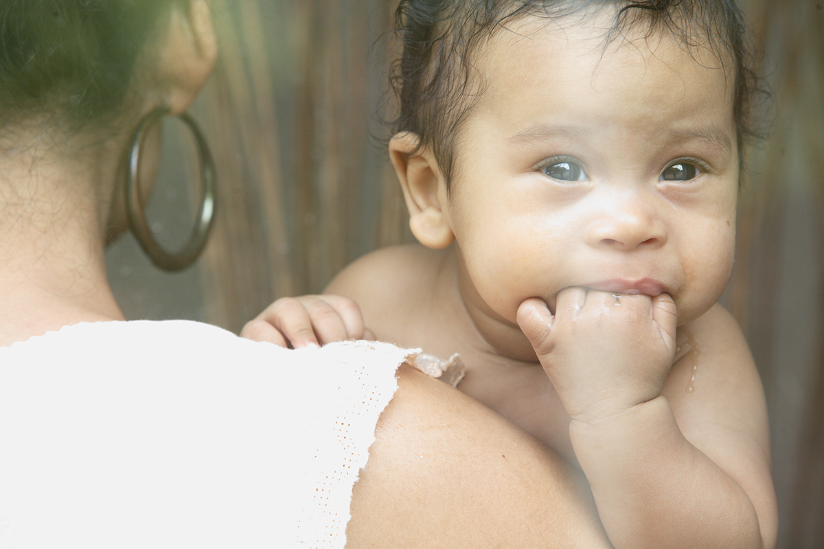 Babyportrait auf Mamas Schulter durch eine Scheibe fotografiert - das baby kaut sabbernd auf seiner Faust herum